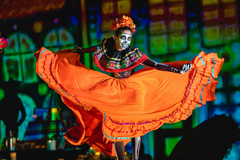 1. MAIN IMAGE_OakBallet-LunaMexicana_VO21469 (1) Smiling female dancer in a bright, flowing orange dress and skull makeup swishing her skirt.